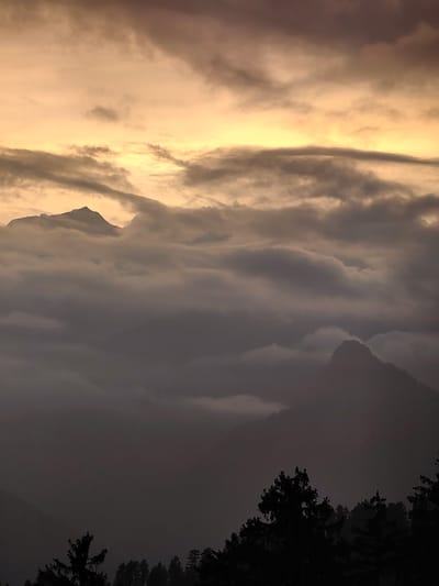 The image depicts a mountainous landscape shrouded in clouds at dusk or dawn. The sky is a gradient of warm, peachy tones at the top, transitioning to darker, more muted grays and purples lower down. Wispy, elongated clouds stretch across the horizon, some tinged with the golden light. In the mid-ground, several mountain peaks are visible, silhouetted against the cloudy sky and lower cloud formations. These peaks appear to be partially obscured by a dense layer of fog or low-lying clouds that blankets the valleys. The foreground is dominated by the dark silhouettes of pine trees, indicating a forested mountainous region. The overall mood is serene and somewhat dramatic due to the lighting and atmospheric conditions. The location is identified as Aleo, India.