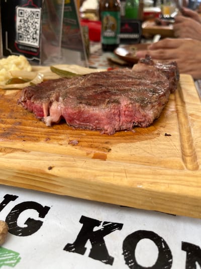 The image captures a close-up view of a piece of meat served on a wooden cutting board. The meat appears to be cooked, with a reddish-pink center and a browned exterior. It sits alongside some pickled vegetables and mashed potatoes. The wooden board shows signs of use, with knife marks and oily residue. In the background, a person's hands are visible, suggesting someone is preparing or serving the food. A QR code is also visible in the background. The environment seems to be an indoor setting, possibly a restaurant, though the specific location within San Francisco Totimehuacán, Mexico, cannot be determined from the image alone. A sign is visible with part of the word "KONG".
