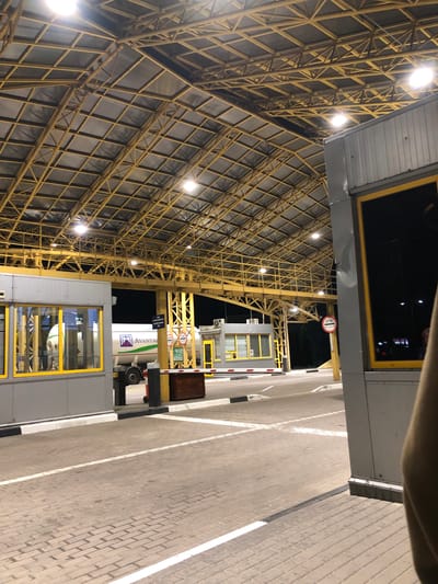 The image captures a nighttime view of a border crossing checkpoint in Starovoitove, Ukraine. A large, yellow metal structure with a complex lattice roof covers the entire scene, illuminated by overhead lights. On the left, a grey booth with yellow-trimmed windows is visible, and a white tanker truck with the "Avantag" logo is parked behind it. To the right, another grey structure with a yellow-trimmed window is present. Roadways are paved with brick pavers and marked with white lines. A red and white striped barrier gate is raised, indicating that traffic is permitted to pass. A circular red and white sign with the word "STOP" and Cyrillic text is mounted on a yellow pole. The background is dark, with hints of trees and distant lights. The overall impression is one of an active, well-lit, and functional border control point.