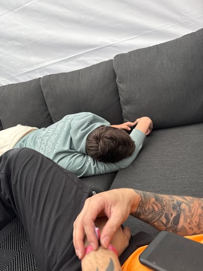 The image shows two individuals sitting on a gray couch in a tent located at 50.442° N, 5.969° E in Stavelot, Belgium. The individual closest to the camera is wearing black pants, an orange shirt, and has a large skull tattoo on their arm. The individual in the background is wearing a light blue hoodie with white lettering and white pants. This individual is lying down on the couch, resting their head on the armrest with their left arm extended. They are holding a black cell phone in their hand. The background of the image is a white tent. The image is taken from a low angle. The lighting is bright and well-lit. The image is a close-up of the two individuals.
