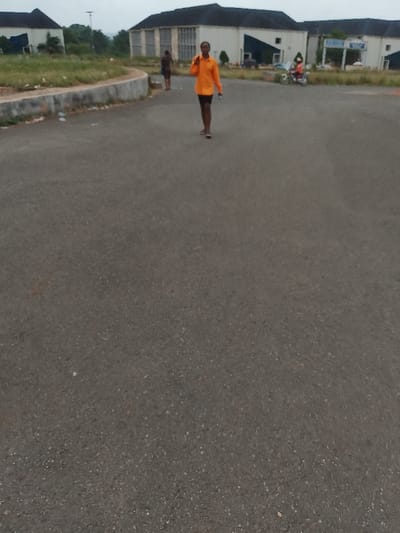 A paved road stretches toward a series of buildings in Ogboko, Nigeria. A person wearing an orange shirt and dark shorts is walking on the road, holding a phone to their ear. A second person is walking behind them to the left. A person on a motorcycle is visible in the background near one of the buildings. The buildings appear to be institutional or public structures. The sky is overcast. Debris and trash are scattered along the edge of the road.