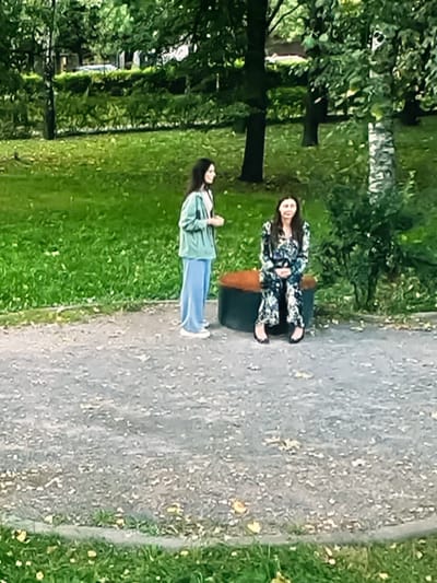 Two women are in a park in Dondușeni, Moldova. The woman on the left is standing and wearing a light blue hooded jacket and loose-fitting blue pants. The woman on the right is sitting on a round, orange and black object, possibly a bench or a planter. She is wearing a long-sleeved dress with a floral pattern. Both women are facing towards the viewer, though the standing woman is looking slightly to her right, and the seated woman is looking directly forward. The park environment includes green grass, trees with dense foliage, and a gravel path. The lighting suggests it is daytime.