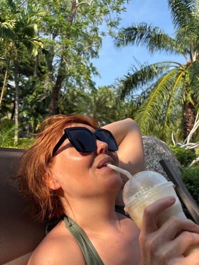 A woman is lying on a mesh sunbed, drinking from a plastic cup with a straw. She is wearing sunglasses and a green bathing suit. The background includes palm trees and other greenery under a blue sky. The location is Khok Kloi, Thailand.
