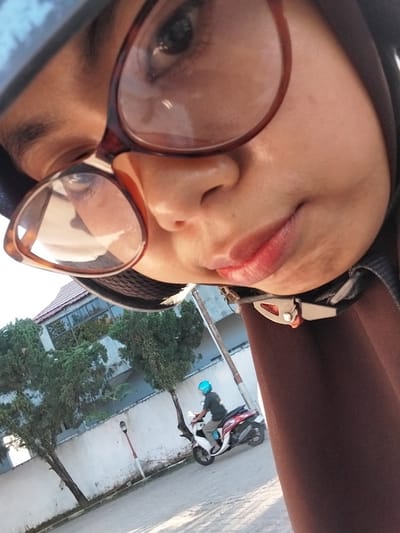 This is a close-up, tilted selfie of a person wearing a brown head covering and large, reddish-brown framed glasses. The person's face is partially obscured by the glasses and the angle of the shot, but their lips, nose, and one eye are visible. Their lips appear to be painted with a pink or red lipstick. The background shows a street scene with trees, a building with a tiled roof, and a motorcycle with a rider wearing a blue helmet. The lighting suggests it is daytime, possibly late afternoon or early morning given the warm tones. The image appears to have been taken outdoors in an urban or suburban setting, with lush green trees indicating a tropical or subtropical climate. The overall mood is casual and personal, typical of a selfie. There is no visible text in the image.