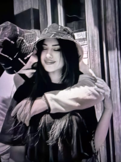 In a dimly lit, outdoor setting with vertical wooden slats in the background, a woman wearing a bucket hat and dark clothing is embraced by another person. The woman's eyes are closed, and she appears to be smiling. Her hair is long and dark. The other person, also wearing a bucket hat with a visible pattern of text, has their arm wrapped around the woman's shoulders. The image quality is grainy and has a purplish tint. There is no indication of the location being Tomai, Moldova.