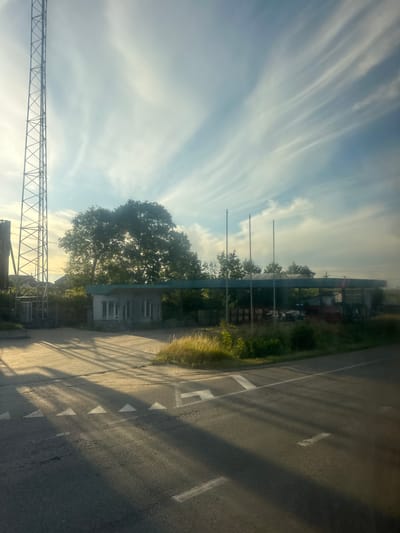 The image depicts a rural scene in Pidbirtsi, Ukraine, on a sunny day with wispy clouds. In the foreground, a paved road with white directional arrows and lane markings is visible, casting long shadows from an unseen object. To the left, a tall, lattice-like transmission tower stands prominently. Further back, a small, single-story building with multiple windows is situated under a covered structure supported by poles. This structure appears to be an abandoned or disused gas station canopy. Behind the canopy, dense green trees and vegetation form a natural backdrop. Several industrial containers and some debris are visible under the canopy. The overall atmosphere is one of quiet, rural isolation, bathed in the warm light of the sun.