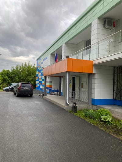 The image depicts the exterior of a building in Danilov, Russia. The building has a modern design with white and gray panels, accented by orange and blue sections. A Russian flag is visible on a pole. The building facade also features a pattern of blue, orange, and green tiles. A black SUV, with license plate A 884 TP 76, is parked on the wet asphalt road in front of the building. Vegetation and trees are seen in the background, and the sky is cloudy.