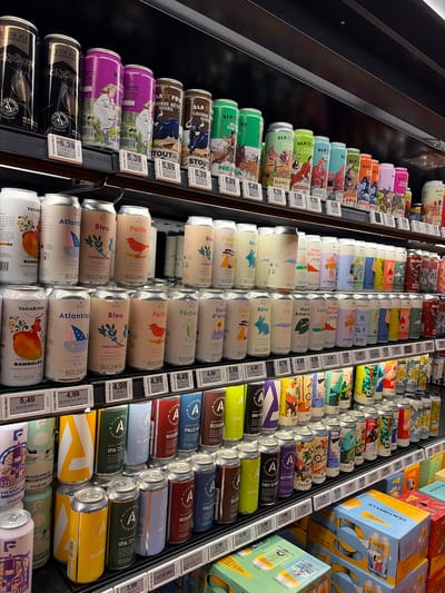 This image captures a vibrant display of various canned beverages, primarily craft beers, arranged neatly on multiple refrigerated shelves inside a store, likely in Saint-Bruno-de-Montarville, Canada. The setting is an indoor retail environment with artificial lighting, presenting merchandise for sale.
Numerous colorful cans are organized in vertical rows, each featuring distinct, often artistic, labels with illustrations ranging from animals like bulls and birds to abstract designs and landscapes. Brands such as "ALA," "BOLDWIN," "ALCHIMIST MICROBRASSERIE," "VAGABOND," and "SENTINELLE" are prominently visible. Many cans are labeled "BIO," indicating organic products.
The cans display various beer styles, including "IPA ROUGE," "STOUT," "SCOTCH ALE," "NEIPA," "GOSE," "DRY STOUT," "PALE ALE," "BLANCHE FESTIVE," and "LAGER MEXICAINE." Alcohol percentages (alc./vol.) are listed, ranging from 2.9% to 8.1%. The can sizes vary, with many being 473ml and some multipacks of 355ml.
Electronic price tags are affixed to the shelves below each product, showing prices from 3.99 to 6.39. The presence of both French and English text on product labels and descriptions ("série terroir," "gose aux bleuets," "IPA de blé," "collection houblonnée") aligns with the bilingual context of Quebec, Canada. No people or specific activities are depicted; the scene focuses on the static product arrangement. Time of day and weather are not discernible.