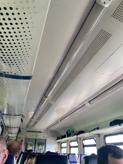 The image shows the interior of a train car, looking upwards towards the ceiling. The ceiling is white and features several light fixtures and ventilation grilles. In the foreground, the heads and shoulders of several passengers are visible, indicating they are seated. On the right side of the image, luggage is stored on an overhead shelf. The windows of the train are visible, revealing a blurry outdoor scene with greenery, suggesting the train is in motion and possibly passing through a rural or suburban area. The overall impression is that of a typical train interior. The location is identified as Kungur, Russia.