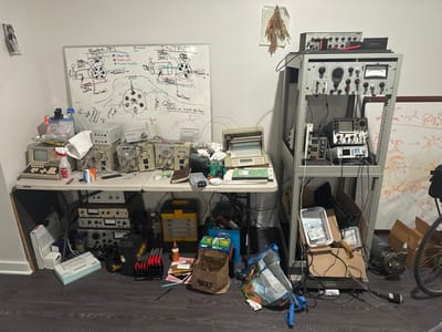 A cluttered workbench features a whiteboard with technical diagrams and notes. Oscilloscopes, vintage computer equipment, and other electronic devices crowd the table and floor space. A metal equipment rack houses more electronics. Below, boxes, bags, and tools add to the disarray. A bicycle is visible on the right side of the frame. The overall impression is of a well-used, functional workspace for electronics and technical experimentation.