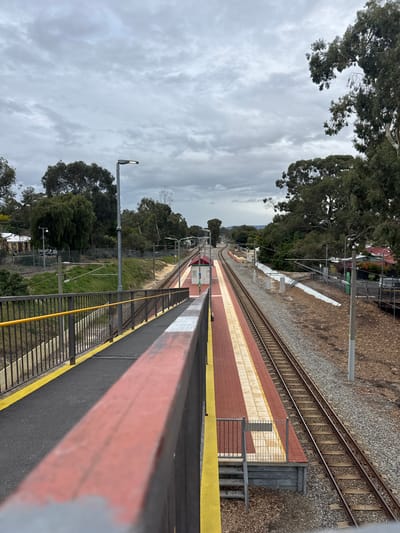 The image depicts a train station platform in Bassendean, Australia, under an overcast sky. The platform is elevated and has a red-and-cream brick surface with yellow safety markings. A black railing with a red top runs along the edge. The train tracks run parallel to the platform. Trees and some buildings are visible beyond the tracks. Streetlights stand on one side of the tracks. The image is taken from a high vantage point.