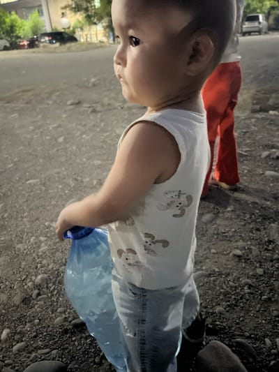 A close-up, side profile shot shows a young child, facing right, wearing a white tank top with cartoon animal prints and light blue denim jeans. The child holds a large, clear blue plastic bottle filled with liquid in their right hand. The child's left arm is bent at the elbow, resting on the bottle. In the background, out of focus, a person wearing red pants stands behind the child. The ground is covered in small pebbles and rocks. The scene appears to be outdoors, possibly on a street or pathway in the city of Şamaldı-Say, Kyrgyzstan, as indicated by the presence of streetlights and buildings in the distance. The lighting suggests it is either dusk or nighttime.