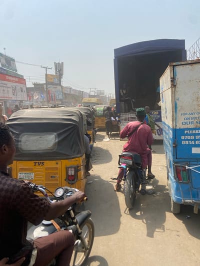 The image depicts a congested street scene in Igbede, Nigeria, under bright, hazy daylight conditions. The foreground features a person on a black motorcycle, observed from the left side, wearing a brown patterned shirt and brown pants. To their right, a queue of yellow TVS-branded Keke Napeps (auto-rickshaws) with black soft tops extends into the mid-ground; the first visible Keke Napep displays a Nigerian license plate "Lagos KJA-890DR". Adjacent to the Keke Napeps, on the right side of the road, a person rides a dark motorcycle, viewed from behind, attired in a maroon tunic and a green cap. This motorcycle has visible blue suspension springs and a partially obscured license plate reading "Lagos F668".
Further along the road on the right, a large blue box truck is parked with its rear door open, revealing at least two individuals within; one person is seated, wearing a striped track jacket. Immediately to the right of the blue truck is the rear-left section of a light blue and white box truck or delivery vehicle, showing some rust and partial text including phone numbers. The road surface appears unpaved and dusty. The background reveals a dense commercial area with numerous buildings, utility poles, overhead wires, and various advertisements for brands such as "FIRMAN", "Airtel", "STAR TIMES", "Legend Extra Stout", and "GLO". A communication tower or mast is visible above the buildings in the distance. Additional vehicles, including more yellow auto-rickshaws, are present in the far background, indicating heavy traffic.