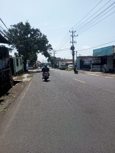 The image presents a daytime view of a paved two-lane road within the city of Beteng_Pangkhar_Somthrang, Bhutan, under a clear blue sky with minimal cloud cover.
In the immediate foreground, a dark-colored motorcycle is observed traveling away from the camera in the left lane. The operator wears a blue helmet and dark clothing. The motorcycle displays a white license plate with partially visible blue characters. A yellow dashed line marks the center of the asphalt road.
To the left of the road, a low stone wall with a dark metal fence runs parallel to the pavement, succeeded by a light-colored building and a large deciduous tree with dense green foliage extending across the upper left frame. The unpaved roadside consists of dirt and sparse grass.
On the right side of the road, a second motorcycle with a rider is visible further down the street, also moving away from the viewer. Multiple utility poles equipped with numerous overhead power lines line this side of the road. A prominent light blue building with a dark awning is situated beyond the utility poles, featuring a white sign displaying dark text, including "MIE AYAM BAKSO" and "TEH GELAS". In the distant background, additional structures, a light green truck, and several cars are present, indicating a populated area. No direct interactions between visible individuals are occurring.