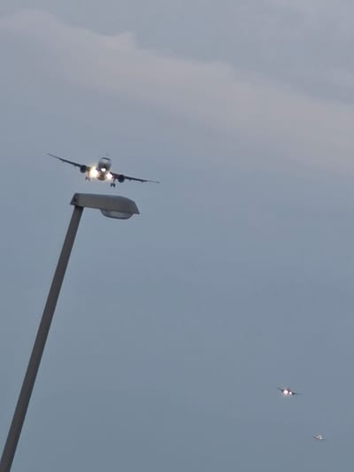 The image captures a scene at Frankfurt Airport (FRA) near Kelsterbach, Germany. In the foreground, a grey lamppost stands at an angle, its light fixture obscured by the sky. In the mid-ground, a large airplane is descending, its landing lights brightly illuminated. Further in the background, two smaller aircraft are visible, also with lights on, suggesting they are also in the process of landing or taking off. The sky is overcast with soft, diffused light, typical of twilight or cloudy conditions. The overall impression is one of an active airport environment with multiple aircraft in the sky.
