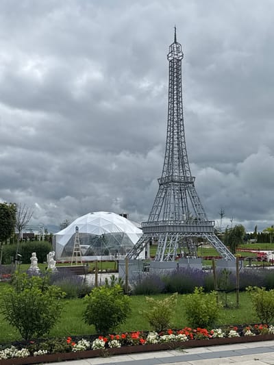 The image captures an outdoor scene featuring a miniature Eiffel Tower and a geodesic dome structure. The Eiffel Tower replica is a metallic, lattice-like structure with intricate detailing, standing prominently in the center-right of the frame. To its left is a large, white geodesic dome, partially covered by a translucent material, with a ladder leaning against it. The foreground is a well-maintained garden with green grass, hedges, and flowerbeds containing red and white flowers. Two white statues of figures, possibly mythological or decorative, are placed in the garden. The background shows more greenery, trees, and a cloudy, overcast sky. The scene appears to be in a park or an amusement area, identified as being located in Bruzdeilynas, Lithuania. There are no people visible in the image.