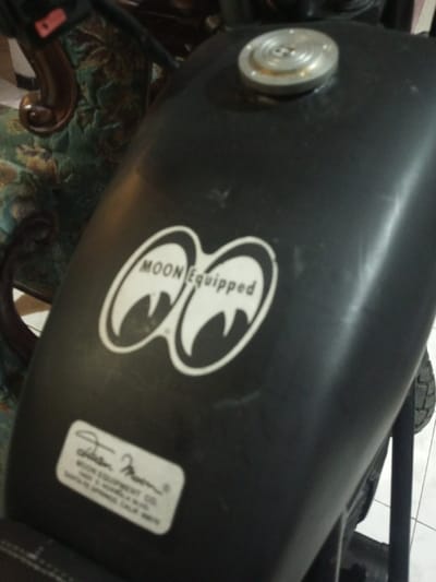 A close-up, slightly overhead view captures the dark-colored fuel tank of a motorcycle. The tank features a prominent white "MOON Equipped" decal, displaying two stylized eye shapes. Below this, a smaller white rectangular decal is visible, bearing what appears to be a signature and the text "MOON EQUIPMENT CO. SANTA FE SPRINGS, CALIF. 90670". A circular, silver-toned fuel cap with concentric ridges is situated on the top surface of the tank. Minor scuff marks and dust are visible on the tank's surface. In the background to the left, a portion of an ornate wooden furniture piece with patterned green upholstery is discernible. To the lower right, a white tiled floor and a section of a motorcycle tire tread are visible. No individuals are present. The scene is located in B. Nam Ngao, Laos.