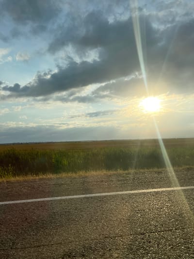 The image depicts a rural roadside scene with a bright sun partially obscured by dramatic clouds. The sun's rays create a lens flare effect, extending across the sky. In the foreground, a dark asphalt road with a white line marking its edge is visible, with a strip of dry grass along the roadside. Beyond the road, a field of tall, green grass extends towards a distant horizon, where the land appears flat with scattered, low-lying vegetation. The sky is a mix of light blue with scattered white clouds and darker, brooding storm clouds. The overall lighting suggests either sunrise or sunset, with a warm, golden hue cast over the landscape. There are no people or animals visible in the image. The scene is a natural landscape with no visible man-made structures other than the road.