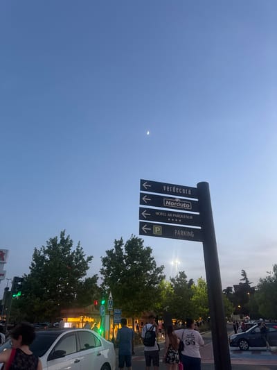 The image captures an evening scene in Leganés, Spain, featuring a directional signpost with directions to "verdecora," "Norauto," "HOTEL AR PARQUESUR," and "PARKING." The sky is a clear blue with a sliver of the moon visible. In the foreground, several people are present, some walking, others standing. A white car is parked on the left side of the image, and other vehicles are visible further down the road. Trees line the background, and traffic lights are illuminated, with one showing green. The overall setting appears to be an urban environment, possibly a commercial area or a street adjacent to a shopping center.
