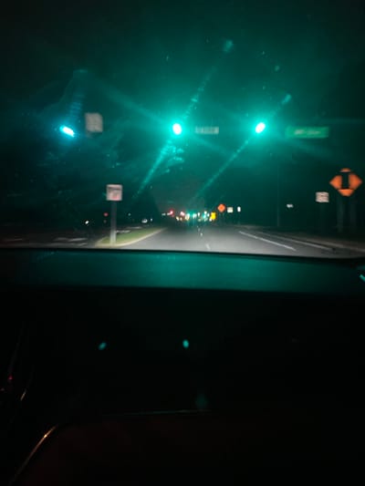 The image depicts a nighttime scene at the intersection of Olden Avenue and Dean Avenue in Lawrence Township, Mercer County, New Jersey, United States. The perspective is from within a stationary vehicle, looking forward through the windshield, with the dark dashboard occupying the lower portion of the frame. Two primary green traffic lights are illuminated overhead, one centered for through traffic or a left turn, and another to its left. A third, smaller green signal is visible on the far left. Overhead street signs identify "OLDEN" (right) and "DEAN" (left). An amber diamond-shaped road sign with a black upward-curving right arrow is visible on the right side of the road. Distant vehicle lights are discernible further down the road. The vehicle's windshield shows visible streaks or smudges. No people are directly visible.