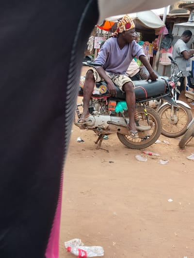 In Ijoko, Nigeria, a person sits astride a motorcycle, their legs dangling on either side. They are wearing a purple long-sleeved shirt and camouflage shorts, with a colorful headscarf tied around their head. The motorcycle is parked on a dirt road, with other motorcycles and market stalls visible in the background. The scene is a typical street view in a Nigerian city, with a bustling atmosphere.