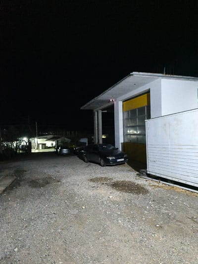 This night scene in Mrzenci, North Macedonia, depicts a gravel and dirt ground surface with several dark, wet patches. To the right, a modern, light-colored building with a grey, flat overhanging roof features a large yellow sectional garage door composed of multiple transparent glass panels. A dark-colored sedan, bearing North Macedonian license plate "GV 0346 LL", is parked directly in front of this garage door. Adjacent to the building on the far right, a white rectangular structure with horizontal corrugations, possibly a trailer or container, is partially visible. In the illuminated background, further to the left, additional buildings and a second, lighter-colored sedan are present. No individuals are visible.
