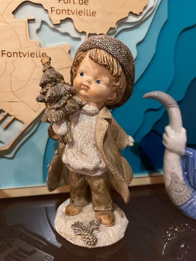 This is a close-up, vertically oriented photograph of a decorative figurine of a young boy. The figurine is made of a ceramic or resin material and is adorned with glitter. The boy is wearing a hat, a coat, and trousers, and he is holding a small Christmas tree. He stands on a circular base. In the background, there is a blue, wavy, layered background that resembles water or stylized waves, and a portion of another figurine or object with a blue garment and a grey arm holding a curved object. To the left of the boy figurine, a portion of a wooden puzzle or map is visible, with the text "Port de Fontvieille" and "Fontvieille" printed on it. The overall scene appears to be a display or collection of decorative items. The lighting suggests it is indoors.
