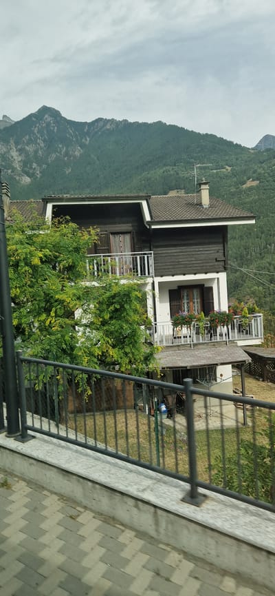 This is a photograph of a two-story house located in Challand-Saint-Victor, Italy. The house has a dark wood exterior on the upper level and white on the lower level. There are balconies with white railings on both floors, adorned with flower planters. A large tree with green foliage partially obscures the left side of the house. In the background, a steep, tree-covered mountain rises under a cloudy sky. The foreground features a black metal fence and a paved walkway. The scene appears to be captured from a slightly elevated position, possibly from a vehicle.