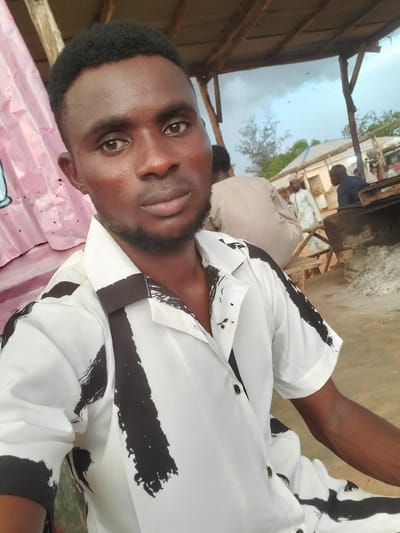 A medium shot shows a young Black man with short, dark hair and a light beard, wearing a white short-sleeved shirt with a black abstract pattern. He is looking at the camera. He is in an outdoor setting that appears to be a marketplace or roadside area in Funtua, Nigeria. In the background, other people are visible, some sitting and some standing, and there are structures that suggest stalls or shops. The sky is cloudy. The man is the primary subject.