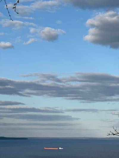 The image shows a body of water, possibly a large lake or sea, under a partly cloudy sky. The sky has a gradient of blue, with some white and gray clouds. The water is dark, reflecting the sky's color. A ship is visible on the water. The location is possibly Galevo, Russia.