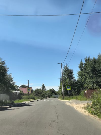 The image depicts a rural road in Afanasyevo, Russia, on a clear, sunny day, likely midday given the bright sky and shadows. The road surface is asphalt with visible cracks and some patches of sand on the right shoulder. A pedestrian crossing is indicated by a yellow square sign with a black pedestrian silhouette, positioned on the right side of the road. Further down the road, another pedestrian crossing sign is visible on the left.
On the left side of the road, there is a fence, followed by a building with a red tiled roof, surrounded by lush green trees and foliage. On the right side, dense green trees and bushes line the road, with a wooden fence in the background. A lamppost with two lights stands near the pedestrian crossing sign on the right. In the distance, a single person appears to be riding a bicycle on the road. Overhead, power lines stretch across the bright blue sky. The overall scene conveys a peaceful, everyday rural atmosphere.