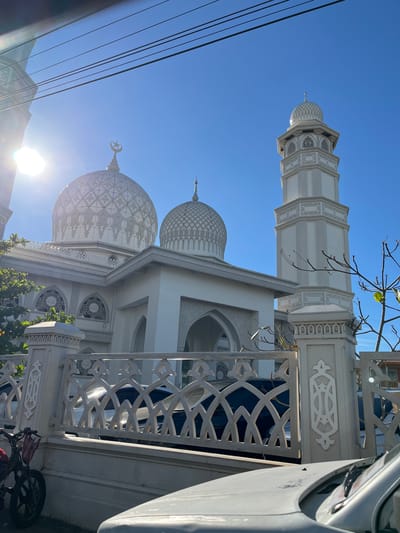 The image captures a mosque with multiple domes and a tall, slender minaret against a clear blue sky. The architecture is predominantly white, featuring intricate geometric patterns on the domes and decorative latticework on the walls and railings. A crescent moon and star symbol adorns the top of one of the smaller domes. In the foreground, a portion of a white vehicle is visible on the right, and power lines are present on the left, suggesting an urban or suburban setting. A bicycle with a red bag is parked near the fence on the right side. The sun is shining brightly, casting lens flare and creating strong highlights on the white structures, indicating it is likely daytime. The scene is serene and appears to be a place of worship in Lambaro, Indonesia. There is no visible text in the image.