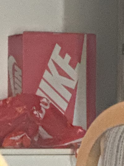 The image shows a close-up of a red shoebox with a white Nike logo. The box is partially obscured by a red object, possibly another shoe or shoe accessory, in the foreground. A person's arm, light-skinned and wearing a grey striped shirt, is visible on the right side of the frame, resting on a light-colored surface. The background is a neutral, textured wall. The scene appears to be indoors, possibly in a home or retail environment. There is no visible indication of the city of Neubrandenburg, Germany.