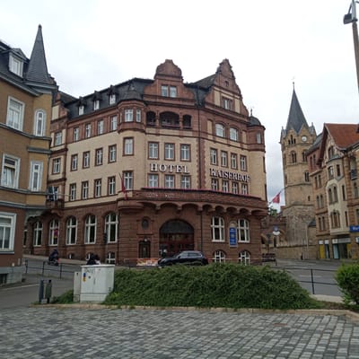 The image depicts a cloudy day in Eisenach, Germany, showcasing the Hotel Kaiserhof and an adjacent building with a prominent church tower. The Hotel Kaiserhof, a substantial multi-story building with a reddish-brown stone facade and lighter colored brickwork, features ornate architectural details, including a curved balcony adorned with flowers. The words "HOTEL" and "KAISERHOF" are clearly visible in white lettering on the facade. A black SUV is parked in front of the hotel entrance.

To the right of the hotel, a tall, slender church tower with a dark pointed roof rises into the sky. The tower has a clock face visible near its peak. The surrounding environment includes a cobblestone plaza in the foreground, a street with traffic and a few pedestrians, and lush green bushes bordering the plaza. A metal trash receptacle is situated on the left side of the street, and a utility box with graffiti is visible near the bushes. In the distance, other buildings with varied architectural styles are present. A motorcycle with a rider is seen on the street, and a woman with dark hair is standing near a parked car in the background.