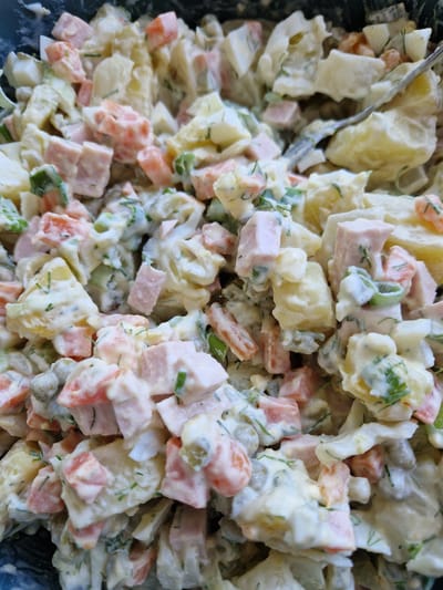 The image is a close-up, overhead shot of a bowl of potato salad. The salad appears to be made with diced potatoes, carrots, peas, ham, and hard-boiled eggs, all mixed with a creamy dressing and garnished with fresh dill. A silver fork is visible on the right side of the bowl, partially submerged in the salad. The background is dark, suggesting the salad is in a bowl or container. There are no people visible in the image, and the setting is a close-up of food, not a specific location in Fresnes, France.
