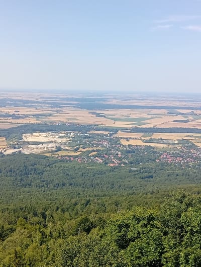 This is a landscape photograph taken from a high vantage point overlooking the city of Sobótka, Poland. In the foreground, a dense forest of green trees fills the bottom portion of the image. Beyond the forest, the city of Sobótka is visible, characterized by a cluster of red-tiled roofs and buildings nestled amongst more trees. Further in the midground, a large, open quarry with pale, layered rock formations is prominent. The background reveals a vast expanse of agricultural fields in various shades of brown and green, stretching towards a hazy horizon under a pale blue sky. The overall scene depicts a rural landscape with urban and industrial elements integrated within it.