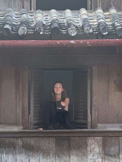 The image shows a woman looking out of a window in a building in Sà Phìn Commune, Vietnam. The building appears to be made of wood and is weathered, suggesting age. The window has a latticework pattern. The woman has fair skin and is wearing dark clothing. Exterior to the window is a column of grey roof tiles. A red pipe runs vertically along the wooden facade next to the tile. The image is taken at an angle.