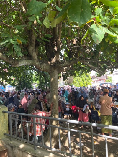 This image depicts a crowded outdoor scene in the city of Pulodolang, Indonesia, under the shade of a large tree. A significant number of people, primarily women dressed in hijabs and modest clothing, are gathered. Some individuals appear to be queuing or waiting, while others are engaged in conversations or observing. Several tables and chairs are visible in the background, some with numbers or labels, suggesting an organized event or distribution. The ground is covered with dirt and scattered leaves. A metal railing runs along the foreground, separating the viewer from the assembled crowd. The overall atmosphere appears to be one of community gathering or a public service event.