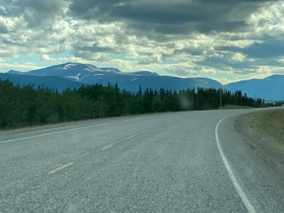 The image depicts a scenic view of a paved road curving through a natural landscape in the vicinity of Mendenhall Landing, Canada. The road, marked with a solid white line and intermittent yellow dashes, appears to be asphalt. On the left side of the road, a dense line of dark green coniferous trees creates a natural barrier, with deciduous trees interspersed. In the background, a range of mountains rises majestically, their peaks dusted with white snow, suggesting a high-altitude or northern climate. The sky is dramatic, filled with heavy, textured gray clouds interspersed with patches of bright blue sky, hinting at dynamic weather conditions. The overall scene is one of an open, undeveloped natural environment, with the road serving as the primary man-made element. There are no people or animals visible in the image.