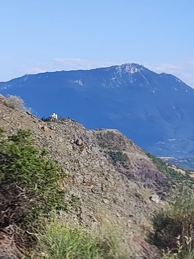 The image depicts a mountainous landscape under a clear blue sky. In the foreground, a rocky, sparsely vegetated slope dominates the scene. Further back, a larger, darker blue mountain range forms the background, with its peak appearing snow-dusted. A small, white building, possibly a church, is situated on a rocky outcrop on the middle ground slope. The overall impression is one of a remote, natural setting. The specific location in Μεγάλη Κερασιά, Greece, is not evident from the visual information provided in the image.