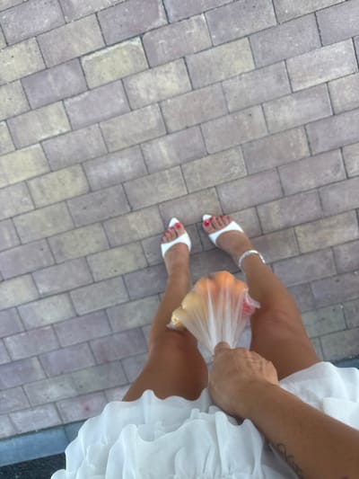 The image depicts a person's legs from a high-angle perspective, standing on a paved surface composed of rectangular bricks in varying shades of gray and tan. The individual is wearing white open-toed heels with bright red painted toenails and a silver anklet. They are holding a clear plastic bag containing what appears to be fruit, possibly oranges and yellow fruits. The person's legs are tanned, and a tattoo is visible on their forearm. They are also wearing a white, ruffled dress or skirt. The scene appears to be outdoors, with the brick pavement suggesting an urban or public outdoor setting. There is no visible indication of the specific location within Zapadny, Russia.