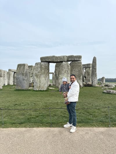 The image displays an outdoor scene at Stonehenge, located near Amesbury, Wiltshire, United Kingdom, under an overcast sky. In the foreground, a light-colored paved path is visible, separated from a green grassy area by a low, dark rope barrier supported by slender metal posts.
To the right of the frame, a man, appearing to be of adult age, stands facing slightly left towards the camera, holding a young child, a toddler, in his arms. The man wears a white collared jacket, dark blue jeans, and white sneakers, and exhibits a slight smile. The child is dressed in a light grey knitted beanie hat, a grey button-up cardigan, light brown trousers, and dark shoes, also looking towards the camera.
The central and background features consist of the ancient monument of Stonehenge. Multiple large, weathered grey sarsen stones are arranged in a circular pattern on the green grass, with some vertical stones supporting horizontal lintel stones atop them, forming trilithons. The stones exhibit natural texture and some areas of lichen growth. Beyond the stone circle, flat green fields extend into the distance, with a faint line of trees visible on the far right horizon.