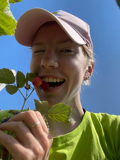 A young person with light hair and freckles is captured in a close-up shot, looking up towards a bright blue sky. They are wearing a light pink baseball cap, tilted slightly, and a vibrant lime green t-shirt. A sprig of raspberries, with two ripe red fruits, is held near their open mouth, with one raspberry touching their teeth. The person is winking with their left eye, and their right eye is partially visible. They have several piercings: a silver ring in their nostril, a small stud on their eyebrow, and multiple hoop earrings in their left ear. A thin silver ring adorns a finger on their left hand. The background consists of a clear blue sky and some blurred green foliage, suggesting an outdoor setting. There is no indication of the specific location within Uva, Russia.