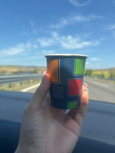 A person's hand, with a ring on the ring finger and pink-tipped fingernails, holds a colorful paper cup adorned with a geometric pattern of squares and dots. The cup contains a small amount of a light-colored beverage. The background is a blurred highway with green fields and trees on either side, under a bright blue sky with scattered white clouds. The scene appears to be captured from inside a vehicle. The image is not specific to the city of Vratitsa, Bulgaria; it depicts a general highway landscape.