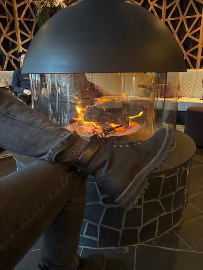 A person wearing black jeans and black boots crosses their legs near a modern fireplace. Flames are visible through the glass of the spherical, partially covered fire pit. Abstract decorative patterns are present on the walls and floor nearby. Reflections of people and objects are visible on the glass surface of the fireplace.