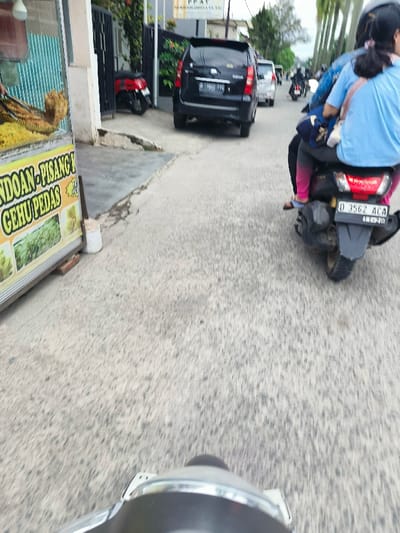The image captures a first-person view from a motorcycle traveling along a street in Kecamatan Dayeuhkolot, Indonesia. The motorcycle is moving forward, and the road is visible ahead. The motorcycle is behind a black scooter with a license plate reading "D 3562 ACA," which is carrying two people. The scooter is followed by several cars, including a black SUV and a white car. The image was taken near a small building with a sign that reads "NDOAN - PISANG GEHU PEDAS." The street appears to be relatively narrow, with buildings and trees visible in the distance. The pavement is gray and slightly rough. The sky is cloudy and overcast. The time of day appears to be afternoon or evening. There are no pedestrians visible in the image. The image is likely captured with a phone or action camera mounted on the motorcycle.