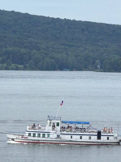 A medium shot shows a two-tiered white passenger boat with a blue canopy on the upper deck, moving left to right across a wide, calm, grey body of water. The boat, named "KARL MARX" in red lettering along its side, has several passengers on both decks. The lower deck has rows of green-tinted rectangular windows. The upper deck is open with railings and passengers standing and sitting. A Russian flag flies from a mast at the front of the boat. In the background, a dense, green, tree-covered hillside rises from the distant shore. A few small buildings are visible on the shoreline amongst the trees. The sky is overcast and a pale grey.