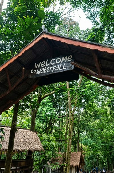 The image captures the entrance to a waterfall in a lush, green forest setting, likely in Dolat Rayat, Indonesia. The focal point is a wooden archway adorned with a weathered sign that reads "WELCOME to Waterfall" in white, hand-painted letters. The archway itself is rustic, with a corrugated metal roof and natural wooden beams.

Beneath the archway, glimpses of traditional thatched-roof huts can be seen, suggesting an established tourist or recreational area. These structures, built with natural materials, blend seamlessly with the dense foliage of the surrounding trees. The forest appears vibrant and healthy, with sunlight filtering through the leaves, indicating it is likely daytime.

While the image is dominated by the natural environment, there are hints of human activity. In the background, several people are visible, appearing to be tourists or visitors, some gathered near one of the huts. Their presence suggests that the waterfall is a destination for recreation and exploration. The overall mood conveyed is one of tranquility and natural beauty, inviting visitors to experience the waterfall. The time of day appears to be late morning or early afternoon, with bright but diffused light. The weather seems pleasant, with no indication of rain or harsh sunlight.