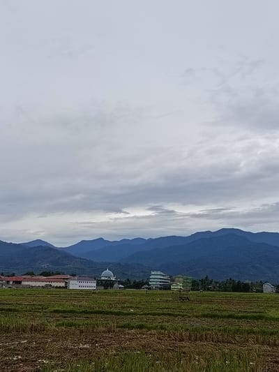 The image depicts a rural landscape in the city of Uleeue, Indonesia. In the foreground, a harvested rice paddy shows stubble and some standing water. Behind the paddy, several buildings are visible. To the left, a long, red-roofed building with white window frames stands adjacent to a white building with multiple floors and numerous windows. In the center, a domed building with a checkered pattern of blue and white, resembling a mosque, is situated amongst other structures. Further to the right, there are more modern buildings, some with green facades, and palm trees. The midground is characterized by a cluster of buildings and vegetation. In the background, a range of dark blue, mist-covered mountains forms the horizon under a cloudy sky. The overall atmosphere is subdued, likely due to overcast weather. No people are clearly discernible in the image.