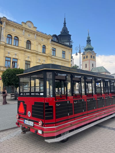 A red and black tourist train, labeled "NEUSOHL EXPRESS," is parked on a cobblestone street in front of a light yellow historic building. The train has multiple carriages with large windows and black accents. A license plate on the back of the train reads "AA 201 YG" and "WWW.AUTOKAVO.SK". A circular sticker with the number "25" is also visible on the train.
The building behind the train features ornate architectural details, including arched windows, decorative carvings, and a clock tower with a green dome. The sky is blue with some scattered white clouds. The scene appears to be outdoors, likely in a city square or historic district. The time of day seems to be late afternoon or early evening, judging by the lighting. The overall impression is one of a pleasant day for sightseeing. A person in a brown outfit is visible near the back of the train.